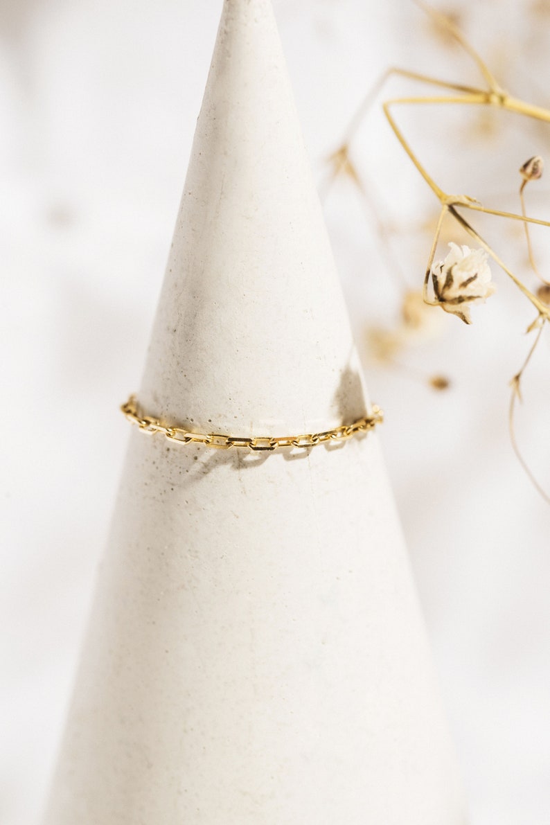 K&ouml;nnte beinhalten: Ein goldenes Kettenarmband mit einem zarten, quadratischen Gliederdesign. Das Armband wird auf einem wei&szlig;en, kegelf&ouml;rmigen St&auml;nder pr&auml;sentiert.