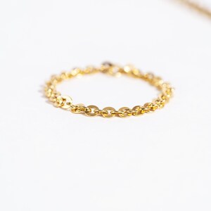 May include: A delicate gold chain ring with a repeating oval link design. The ring is set against a white background, with a blurred gold chain visible in the background. The ring is a simple, elegant piece of jewelry.