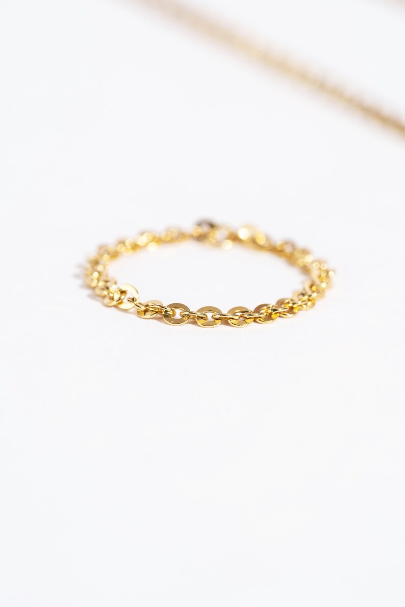 May include: A delicate gold chain ring with a repeating oval link design. The ring is set against a white background, with a blurred gold chain visible in the background. The ring is a simple, elegant piece of jewelry.