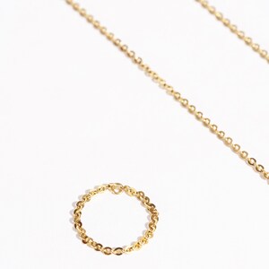 May include: A gold-colored chain ring and necklace on a white background. The ring is a delicate chain design, and the necklace is a longer chain. The jewelry is displayed with a bright, natural light.
