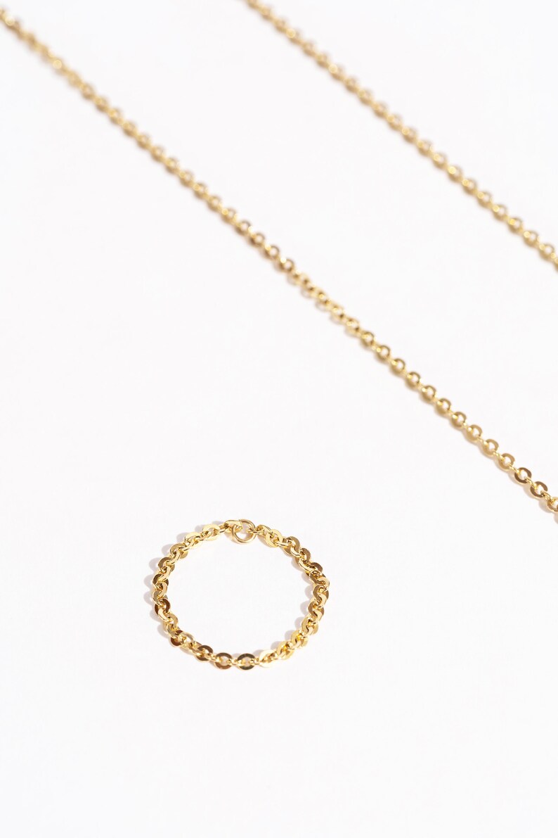 May include: A gold-colored chain ring and necklace on a white background. The ring is a delicate chain design, and the necklace is a longer chain. The jewelry is displayed with a bright, natural light.