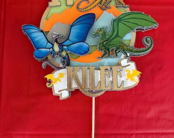 May include: A cake topper featuring a white banner with the name "Kilee" in gold glitter letters. The banner is decorated with two yellow stars. The topper also features four dragons: a green dragon, a blue butterfly dragon, and two orange dragons.