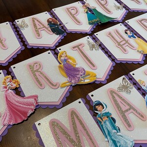 May include: A white and pink banner with glitter letters spelling "Happy Birthday" with Disney princess characters on each letter. The banner has a scalloped edge and is decorated with glitter butterflies.