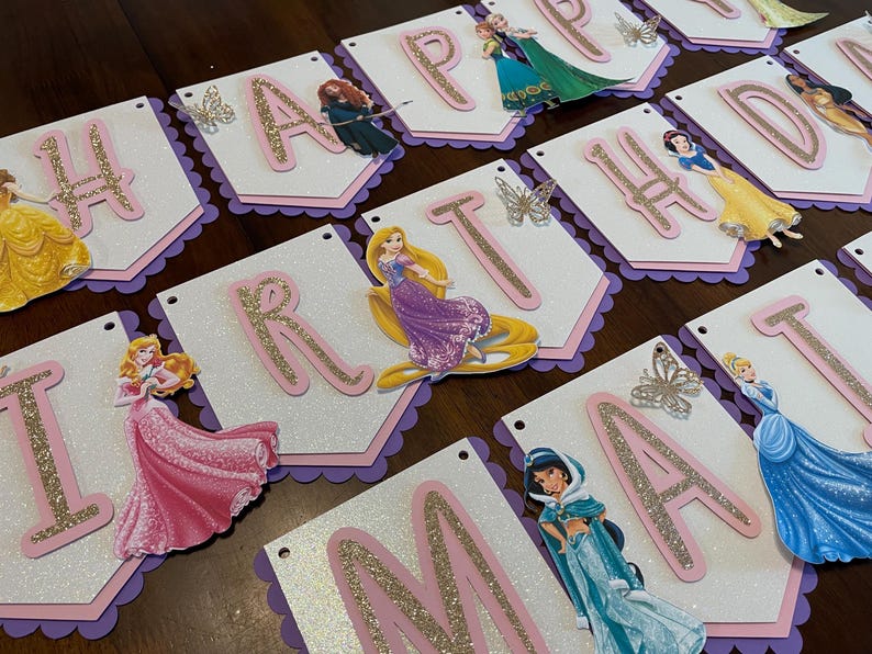 May include: A white and pink banner with glitter letters spelling "Happy Birthday" with Disney princess characters on each letter. The banner has a scalloped edge and is decorated with glitter butterflies.