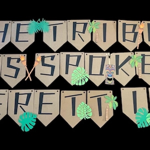 May include: A brown paper banner with the text "The Tribe Has Spoken Everetti Us 12" in black lettering. The banner is decorated with green paper palm leaves and tiki figures.