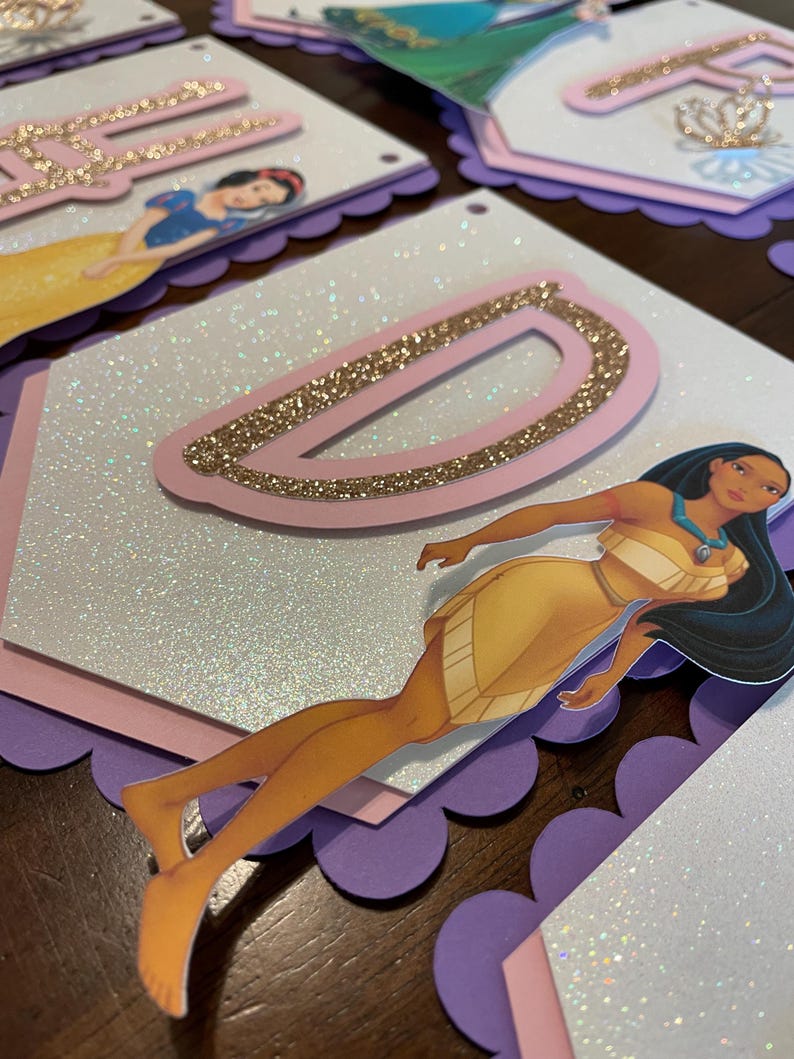 May include: A purple, pink, and white banner with a glitter finish. The banner features a large pink letter "D" with gold glitter accents. A cutout of Pocahontas from Disney's Pocahontas is on the banner.