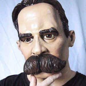 Puede incluir: Primer plano de la cara de un hombre con un bigote marrón oscuro. Lleva una camisa negra y tiene una expresión pensativa.