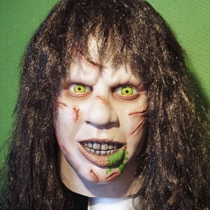 May include: A horror movie mask with long brown hair, a pale face with scars, green eyes, and a green substance on the mouth.