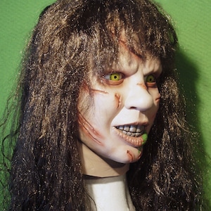 May include: A close-up of a horror movie mask with long brown hair. The mask has a pale complexion with green eyes and a wide, toothy grin. There is green slime dripping from the mouth.