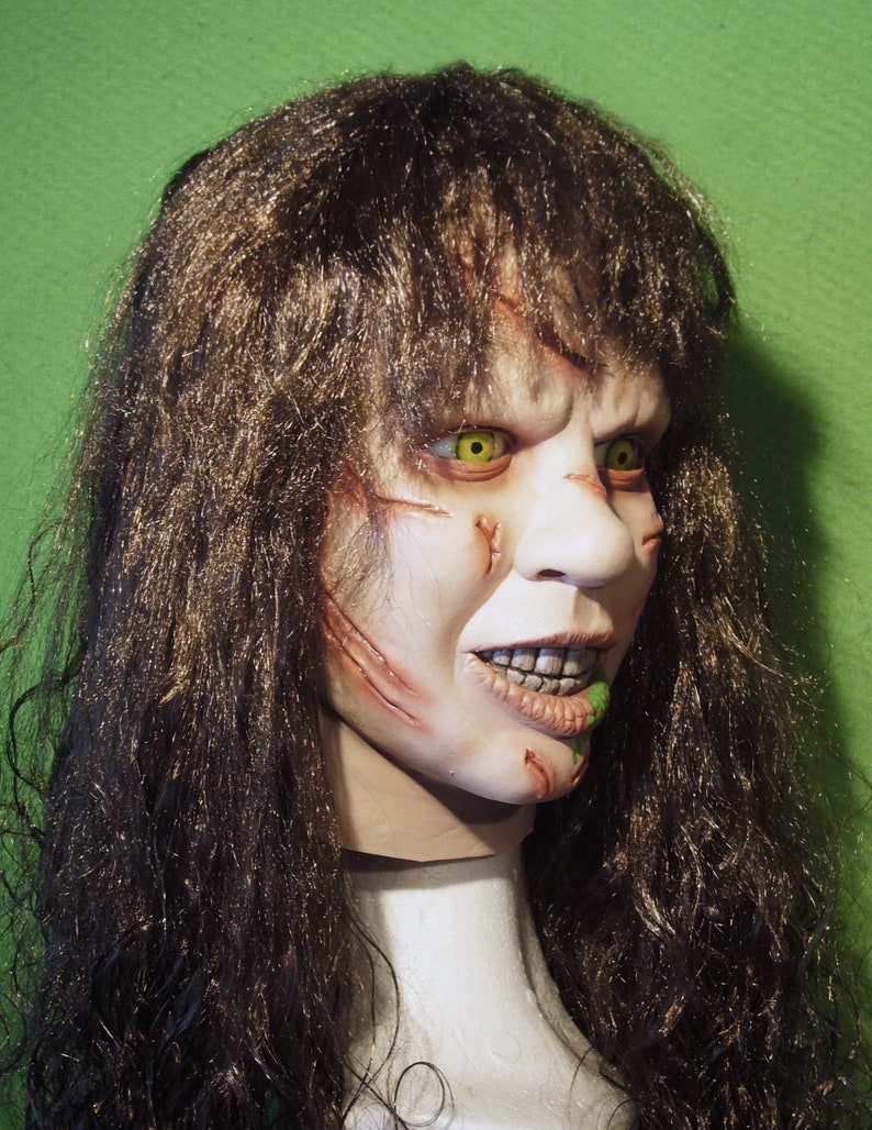 May include: A close-up of a horror movie mask with long brown hair. The mask has a pale complexion with green eyes and a wide, toothy grin. There is green slime dripping from the mouth.