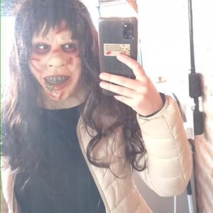 May include: A person wearing a white puffy jacket over a black shirt and blue jeans. They are holding a black smartphone with a sticker on the back. The person is wearing a scary mask with red and white paint and a bloody mouth.