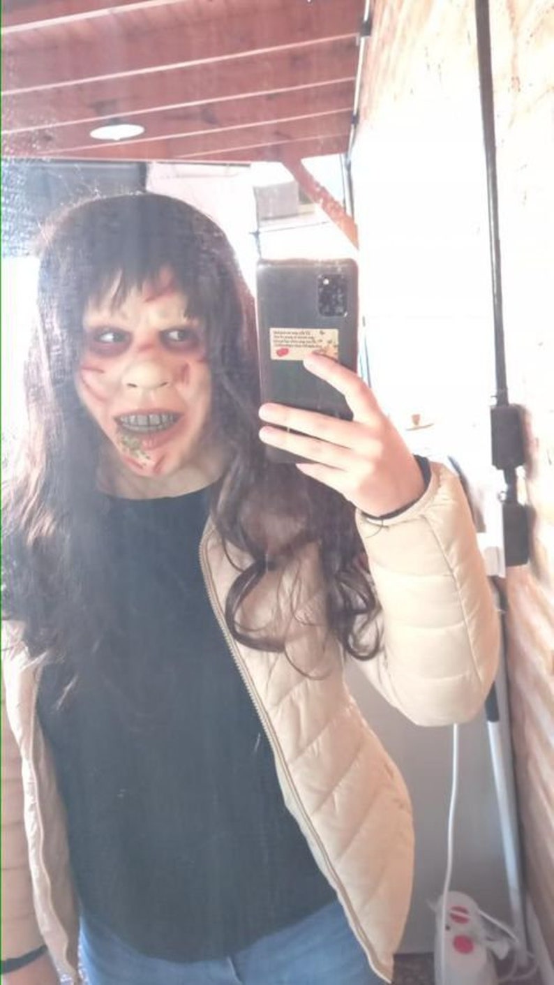 May include: A person wearing a white puffy jacket over a black shirt and blue jeans. They are holding a black smartphone with a sticker on the back. The person is wearing a scary mask with red and white paint and a bloody mouth.