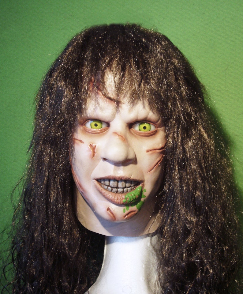 May include: A horror movie mask with long brown hair, a pale face with scars, green eyes, and a green substance on the mouth.
