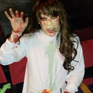May include: A person dressed as a zombie with a white shirt, long brown hair, and red and green makeup. They are holding a glass of a green drink with a straw. The person is sitting in a chair with a red seat and a wooden back.