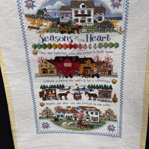 May include: A cross-stitch sampler titled Seasons of the Heart featuring four horizontal scenes of houses, landscapes, and horse-drawn carriages. The embroidery includes text, small metal charms, and pink buttons on a light fabric with a yellow border.