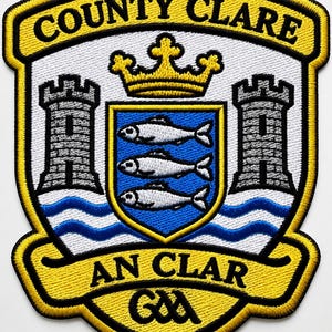 Op de afbeelding: Een geborduurde patch met het wapen van County Clare, een gele rand, witte achtergrond, twee grijze torens, een centraal blauw schild met drie zilveren vissen, een gele kroon en blauwe golven. Tekst: COUNTY CLARE en AN CLAR GAA.