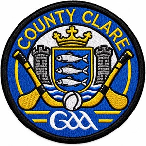 May include: A circular embroidered patch featuring the text COUNTY CLARE in yellow. The design includes a central shield with three white fish, a yellow crown, two grey towers, two crossed yellow hurling sticks, a white ball, and the GAA logo.