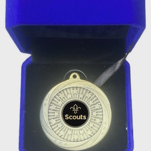 Könnte beinhalten: Goldene Scouts-Medaille mit schwarz-weißem Zentrum, präsentiert in einer offenen blauen Samtbox. Die Medaille zeigt ein Sternmuster und das Wort "Scouts". Ein schwarzes Band ist befestigt.