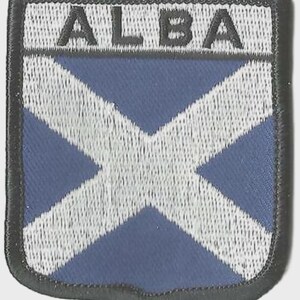 May include: Embroidered patch featuring the word "ALBA" above a white St. Andrew's cross on a blue background. The patch has a black border and a shield shape, suitable for application on clothing or accessories.