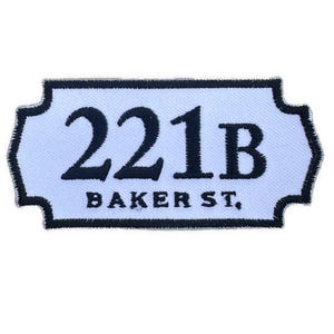 May include: A rectangular patch with a light blue background and a black border. The text "221B BAKER ST," is embroidered in black, centered on the patch. The design is reminiscent of a street sign.