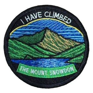 Może przedstawiać: Okrągła, haftowana naszywka z czarną obwódką. Przedstawia górski krajobraz z napisem "I HAVE CLIMBED" u góry i "THE MOUNT SNOWDON" u dołu. Projekt wykorzystuje odcienie zieleni, błękitu i żółci. Idealna dla miłośników gór.