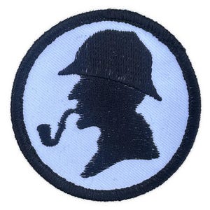 May include: Embroidered patch featuring a black silhouette of a man wearing a hat and smoking a pipe, set against a light blue background. The patch is circular with a black border, suitable for clothing or accessories.