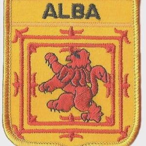 May include: Embroidered patch with a yellow background and a red lion rampant design. The word "ALBA" is stitched in black at the top. The patch has a decorative red border and a gold stitched edge.