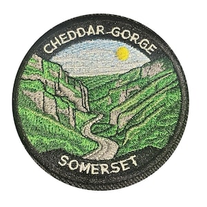 May include: Circular embroidered patch depicting Cheddar Gorge, Somerset. The design shows a winding road through green hills, with the text "CHEDDAR GORGE SOMERSET" around the edge. The patch has a black border and a white background.