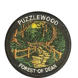 May include: Embroidered patch with a circular design featuring a forest scene. The patch has a black border with the words "PUZZLEWOOD" and "FOREST OF DEAN" in white. The scene depicts a path, trees, and a yellow sky.