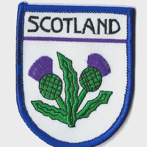 May include: A shield-shaped embroidered patch with a blue border, displaying the word "SCOTLAND" above a thistle emblem. The thistle, in purple and green, is a national symbol. The patch has a white background, suitable for application on clothing or bags.