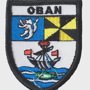 May include: Embroidered patch with a shield design. The top section features the word "OBAN" in black, above a blue square with a white creature and a yellow and black design. The bottom section shows a ship on blue water.
