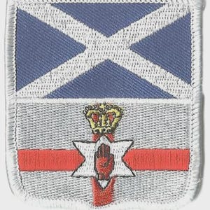 May include: Embroidered patch featuring a blue and white saltire design over a gray background. Below, a red cross intersects a white star with a red hand, topped by a gold crown. The patch has a white border.