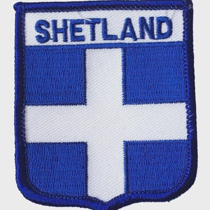 May include: Embroidered patch with the word "SHETLAND" at the top. The design features a white cross on a blue background, with a blue border. The patch is shield-shaped, with a blue stitched edge.