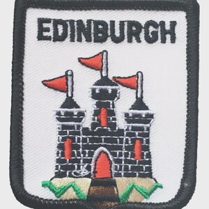 May include: Embroidered patch with the word "EDINBURGH" at the top. The patch depicts a black castle with red flags and accents, set against a white background. The patch has a black border and is a souvenir or emblem.