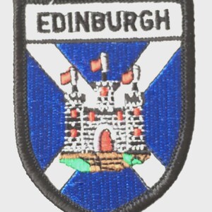 May include: Embroidered patch featuring the word "EDINBURGH" above a blue and white shield design. The shield depicts a castle with red flags and a red arched doorway. The patch has a black border.