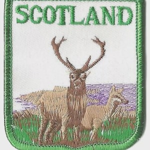 May include: Embroidered patch with the word "SCOTLAND" in green. It features a brown deer and a smaller deer, set against a landscape with grass and water, within a green border.
