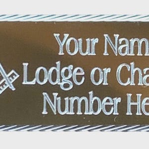 May include: A rectangular name badge with a gold background and a silver border. The badge features the Masonic Square and Compasses symbol and the text "Your Name", "Lodge or Chapter", and "Number Here" in silver lettering.