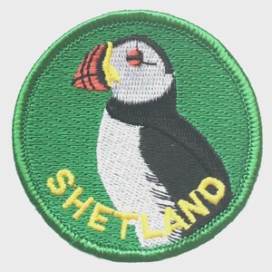 May include: Embroidered patch featuring a puffin bird with a red, yellow, and black beak, and black and white feathers. The patch is circular with a green background and the word "SHETLAND" in yellow.