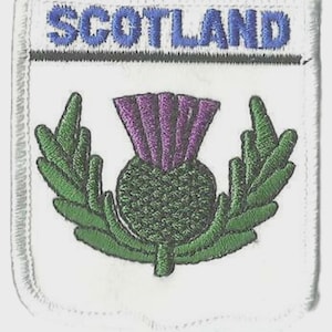 Pode incluir: Patch bordado com a palavra "SCOTLAND" em azul. Apresenta um cardo com flor roxa e folhas verdes, sobre fundo branco com borda costurada.