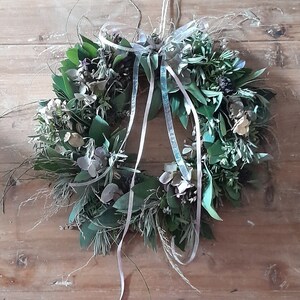 May include: A green and white wreath made of natural foliage and flowers. The wreath is decorated with a silver ribbon tied in a bow.