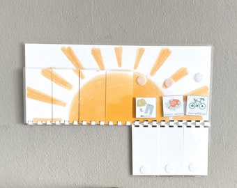 May include: A rectangular visual schedule board featuring a yellow sun illustration with vertical flip panels. Three square cards with icons for clothing, washing, and cycling are attached. Below, three blank white flip panels are mounted on a white plastic spine.