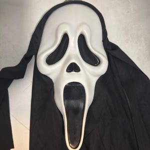 May include: A white plastic mask featuring an elongated face with large, hollow black eye sockets, a small triangular nose, and a wide, open black mouth. The mask is attached to a black fabric hood that drapes down from the top and sides.
