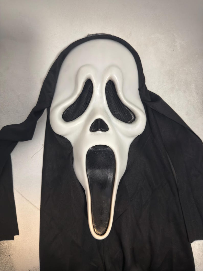 May include: A white plastic mask featuring an elongated face with large, hollow black eye sockets, a small triangular nose, and a wide, open black mouth. The mask is attached to a black fabric hood that drapes down from the top and sides.