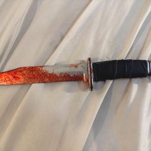 Puede incluir: Un cuchillo de caza con una hoja plateada manchada de rojo. El mango está envuelto en material negro y tiene un extremo color cobre. El cuchillo está sobre una superficie blanca.