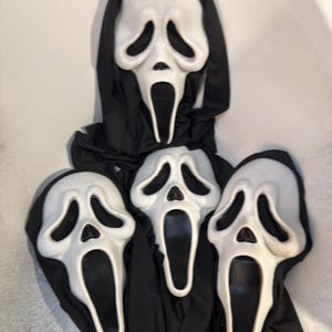 May include: Three white plastic masks with elongated, distorted facial features, including hollow black eye sockets and an open, oval-shaped mouth. Each mask is attached to a black fabric hood that drapes downward, covering the head and neck area.
