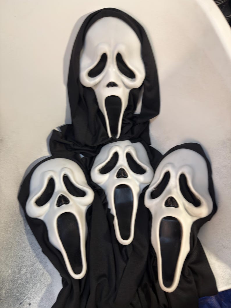 May include: Three white plastic masks with elongated, distorted facial features, including hollow black eye sockets and an open, oval-shaped mouth. Each mask is attached to a black fabric hood that drapes downward, covering the head and neck area.