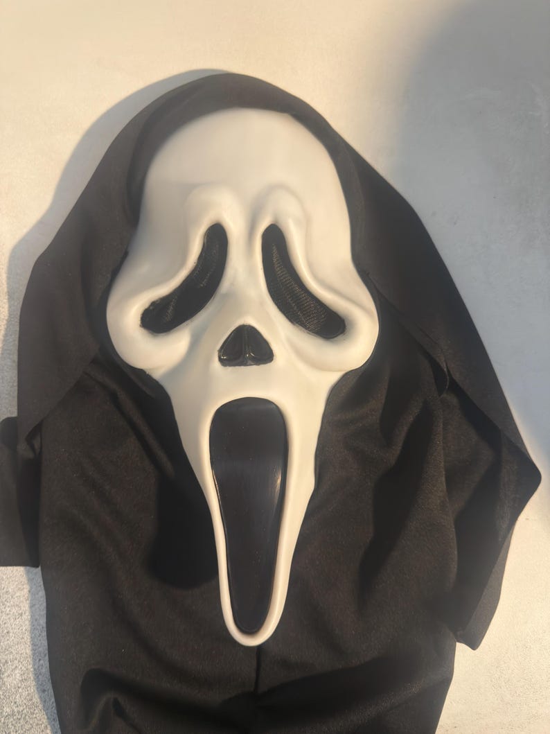 May include: A white plastic ghost mask with elongated black eye sockets and a wide, open black mouth. The mask is attached to a black fabric hood that drapes around the sides and top, creating a full-face covering with a hollow, distorted expression.