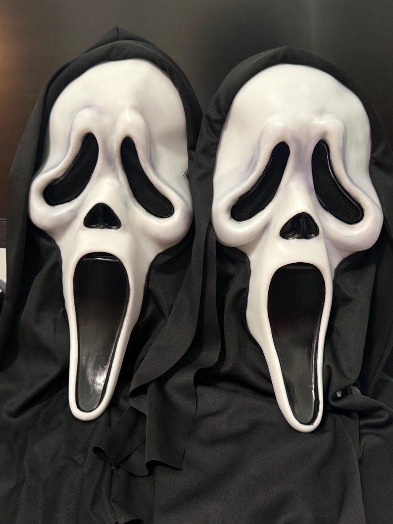 May include: Two white plastic ghost masks with elongated black eye sockets, a small triangular nose, and a wide, open black mouth. Each mask is attached to a black fabric hood that drapes down, covering the sides and creating a dark, uniform appearance.