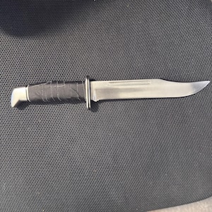 May include: A silver hunting knife with a black wrapped handle and a silver bolster. The blade is long and curved, with a sharp point. The knife is lying on a dark surface.