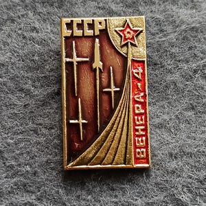 May include: A rectangular vintage pin with gold trim and a red and brown background. The pin features the text "CCCP" at the top, with a red star and the text "ВЕНЕРА-4" on the right side. The pin depicts rockets and a stylized launch.